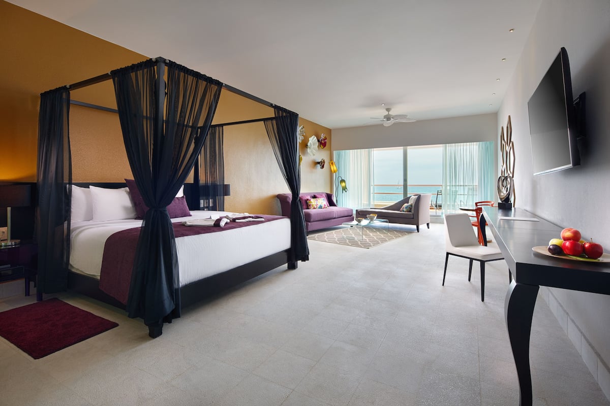 Ein modernes Hotelzimmer mit Himmelbett, einer Sitzecke, einem Esstisch mit Obst und einem großen Fenster mit Blick.