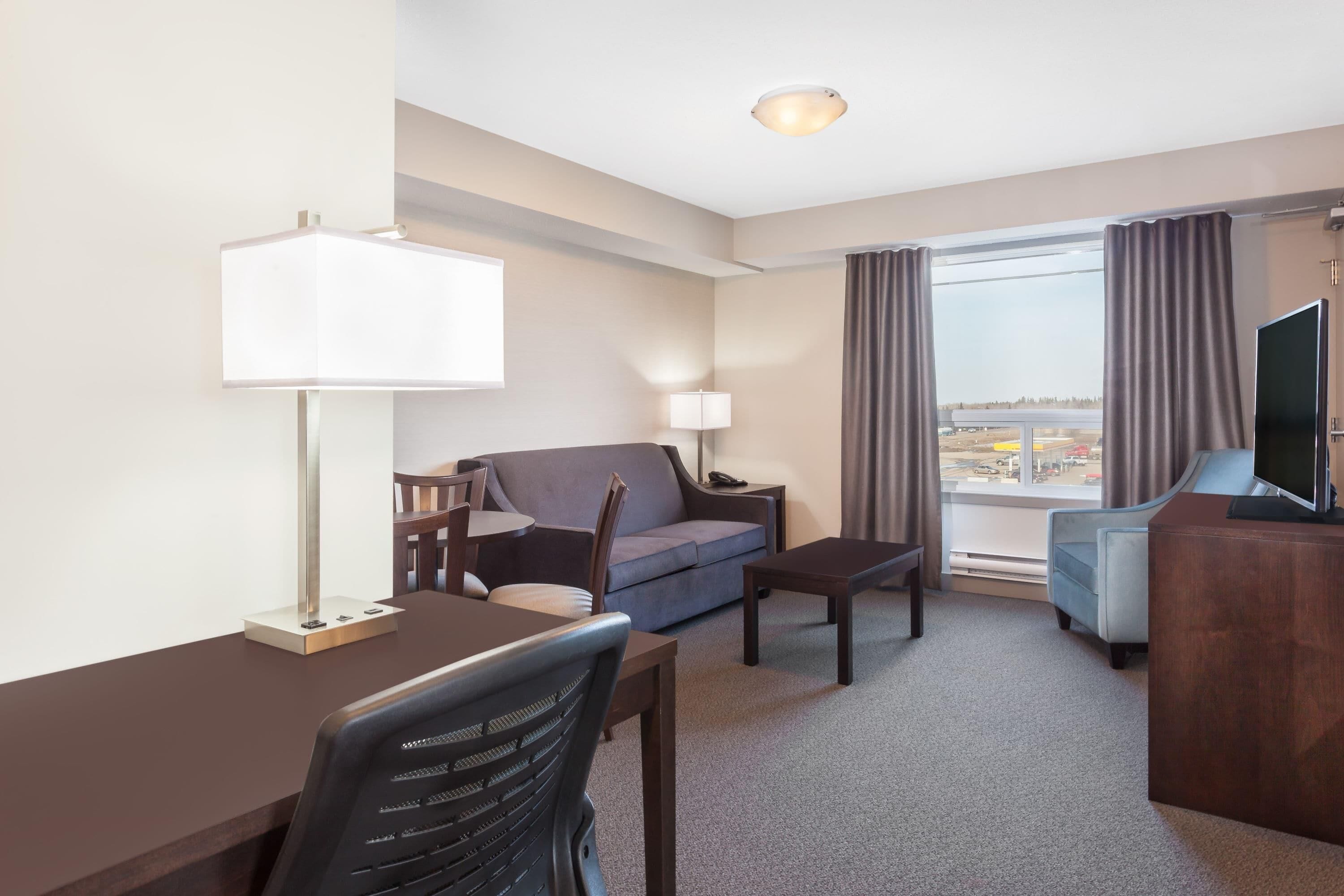 A hotel room with a desk, chair, sofa, TV, and a view of the outside.
