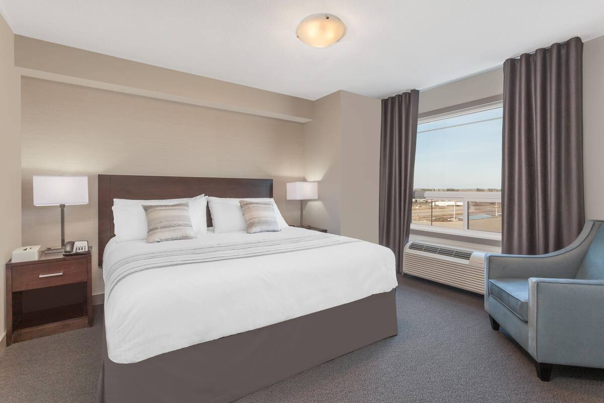 Una habitación de hotel con una cama grande, lámparas de noche, una silla, un ventanal con cortinas y una luz en el techo.