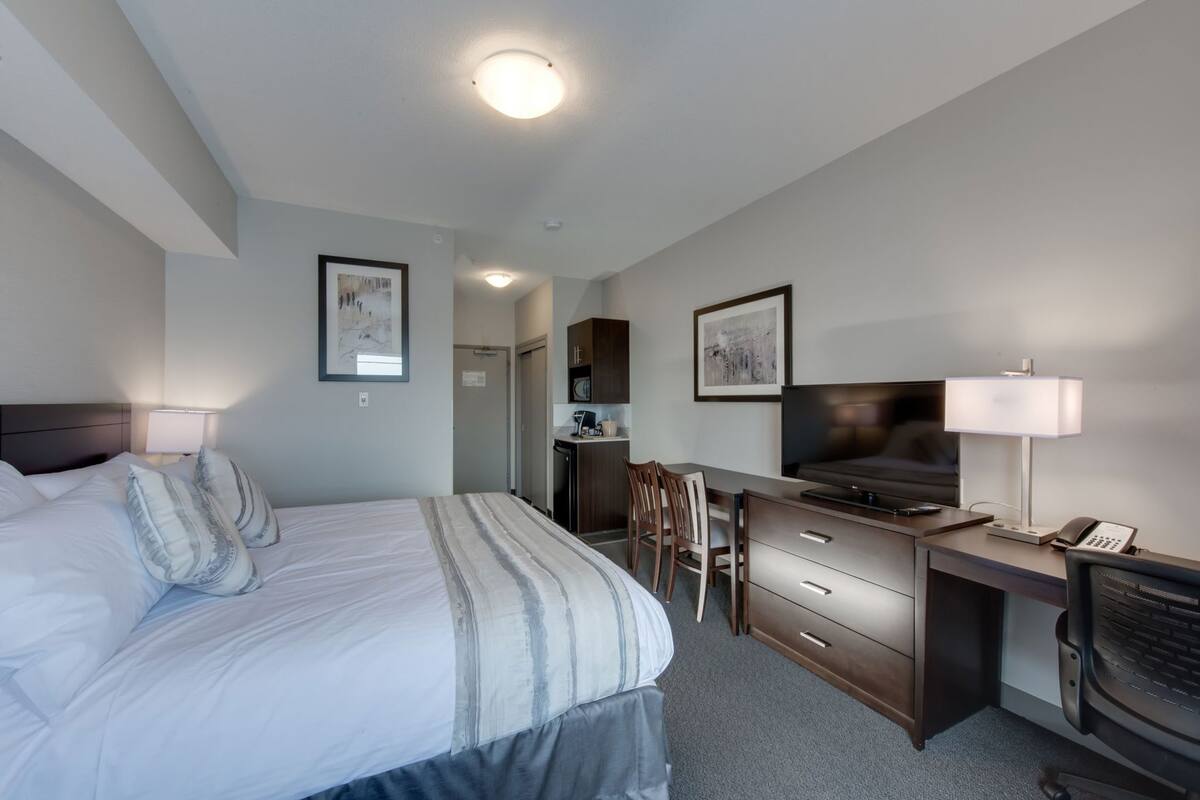 Habitación de hotel con cama, escritorio, televisor, lámpara y una pequeña kitchenette.