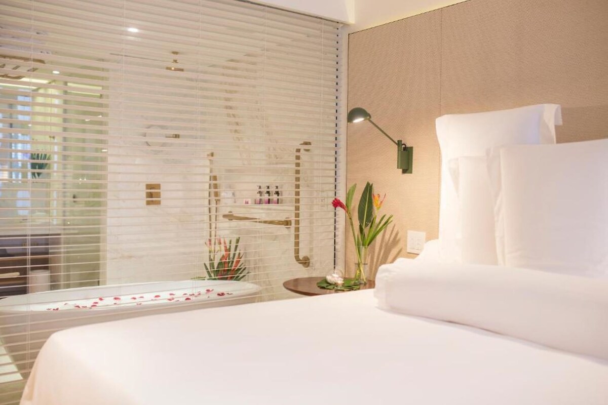 A modern hotel room with a large bed, a bedside table, a lamp, and a window with blinds.
