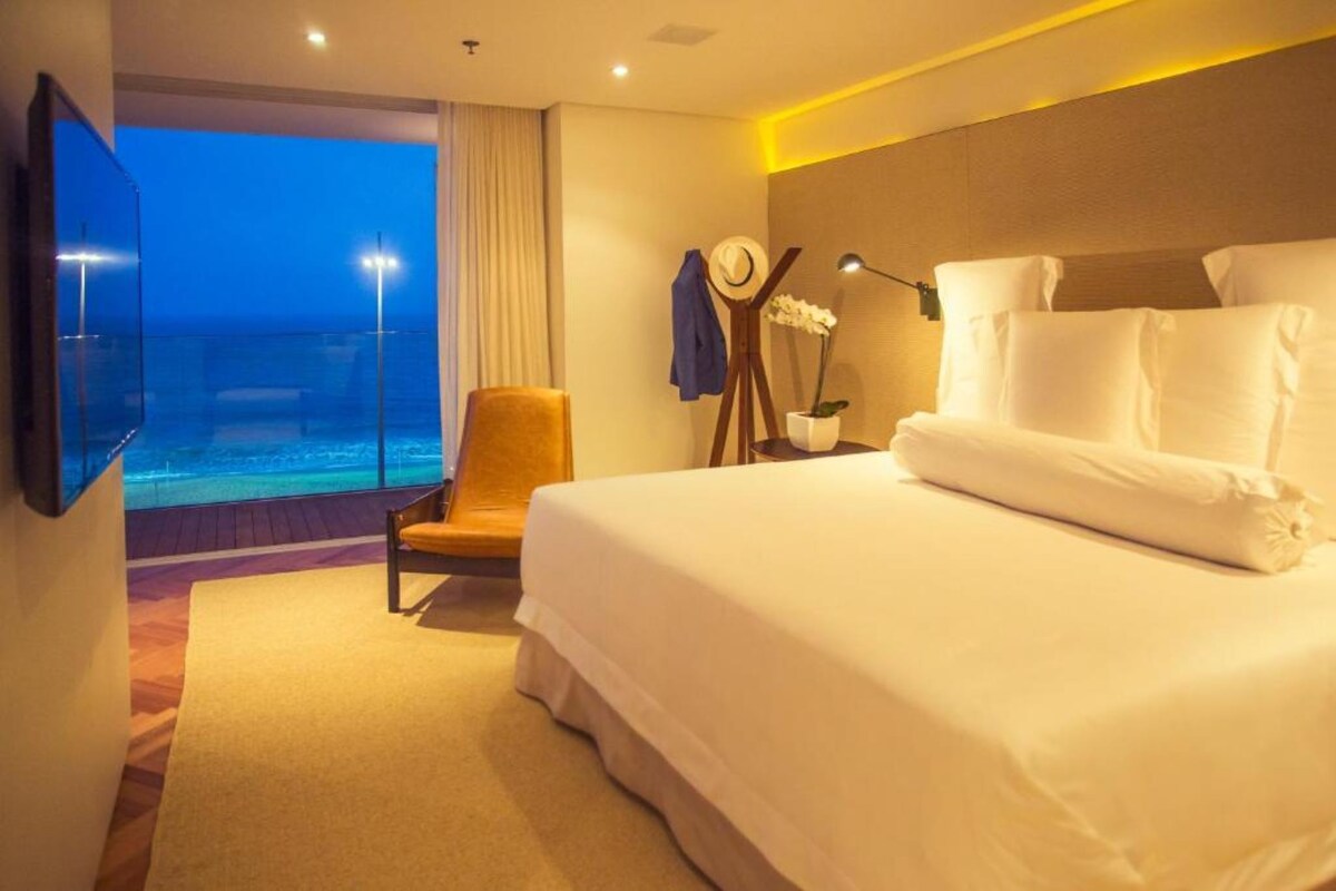 A hotel room with a large bed, a chair, a TV, a nightstand, and a view of the ocean.