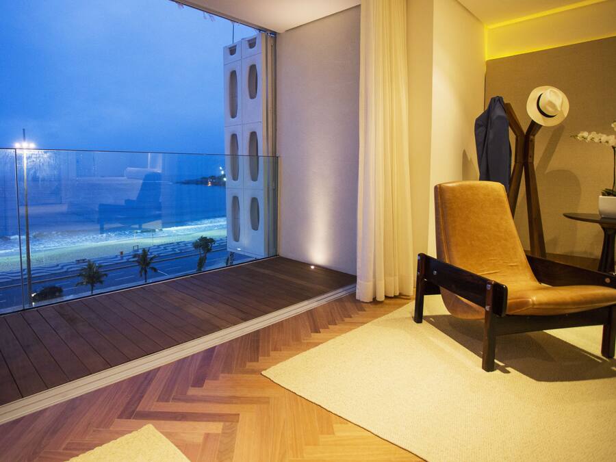 A modern room with a large glass balcony overlooking a beach, a wooden floor, and a comfortable chair.