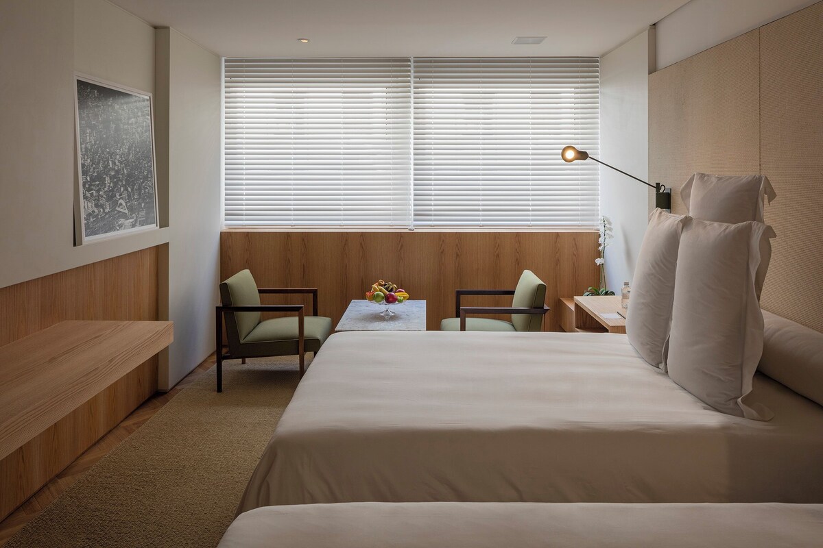 A hotel room with a bed, two chairs, a small table with a vase, and a window with blinds.