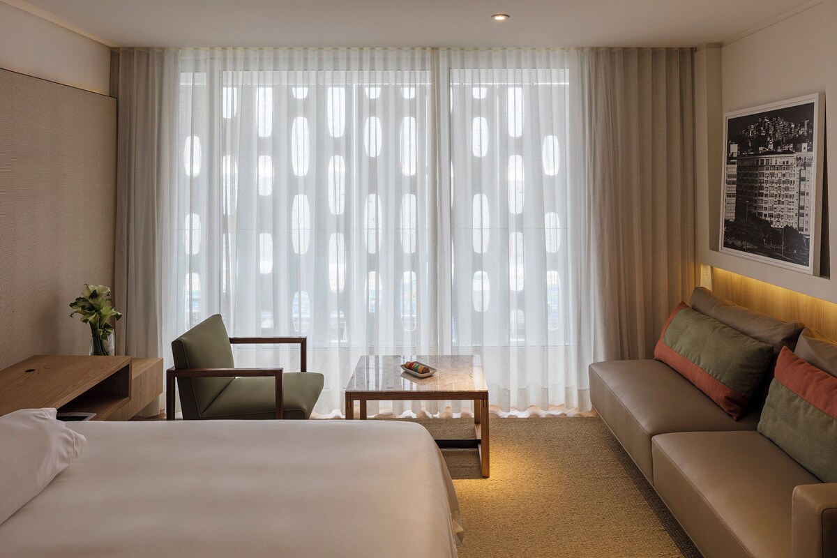 A hotel room with a bed, a sofa, a small table, a chair, and a large window with sheer curtains.