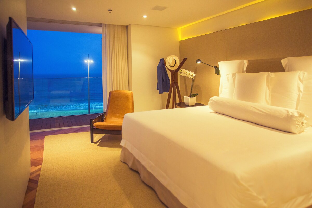 A hotel room with a large bed, a chair, a TV, and a view of the ocean.