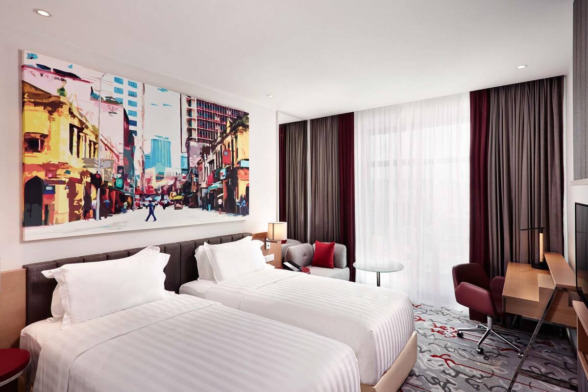 A hotel room with two beds, a desk, a chair, a large painting, and a view of the city.