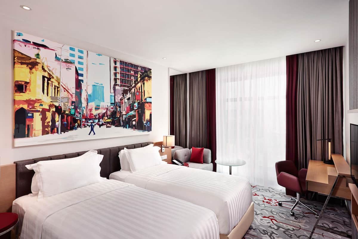 A hotel room with two beds, a desk, a chair, a large painting, and a view of the city.
