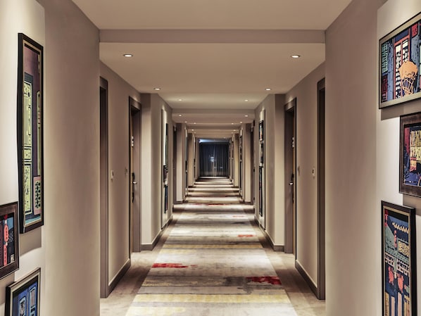 Ein langer, schmaler Hotelgang mit Türen an beiden Seiten, einem gemusterten Teppich und gerahmten Bildern an den Wänden.