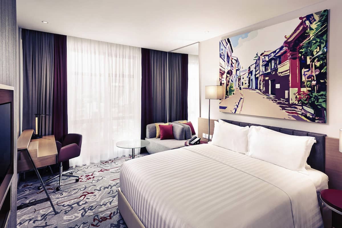 A hotel room with a large bed, a desk, a chair, a small table, and a painting on the wall.
