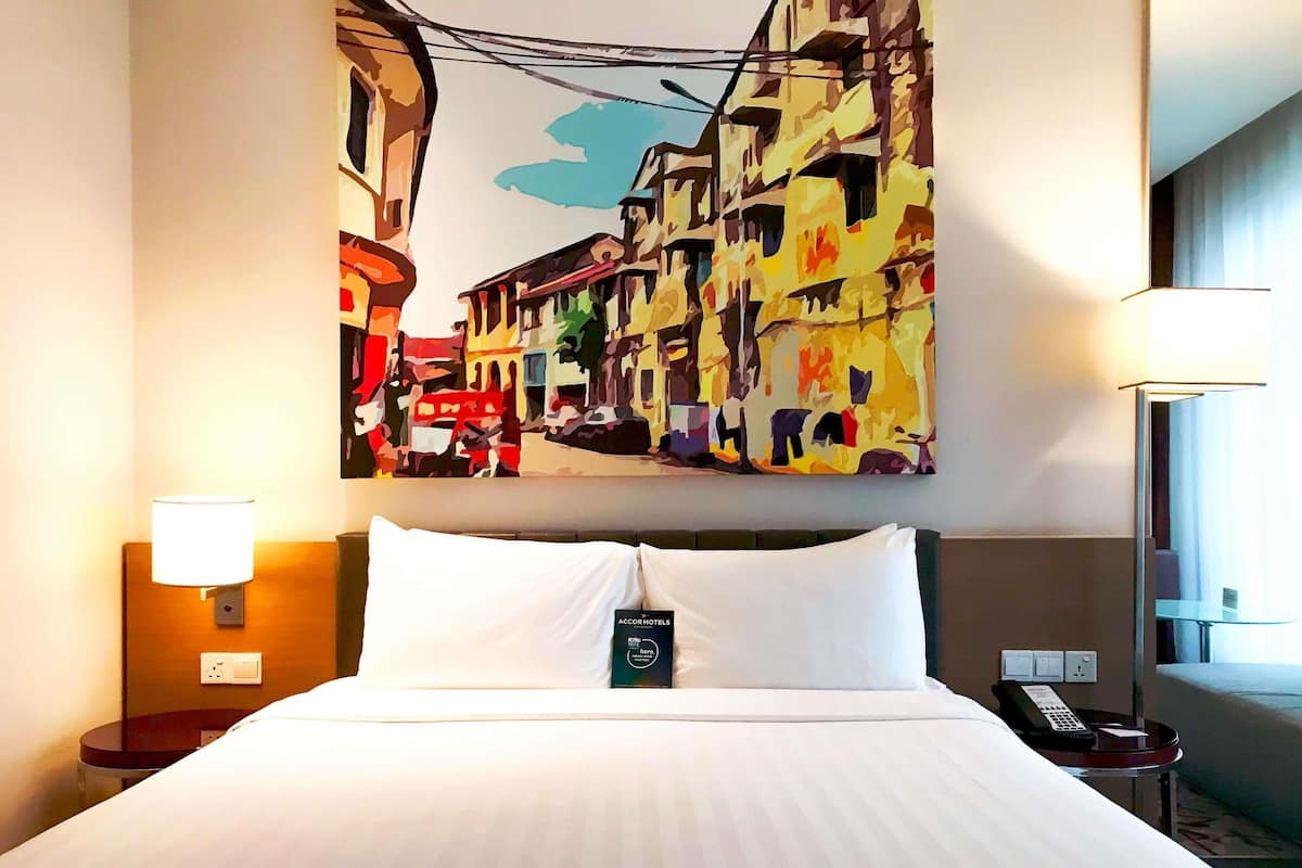 A hotel room with a large bed, a nightstand, a lamp, a painting of a street scene, and a phone on the nightstand.