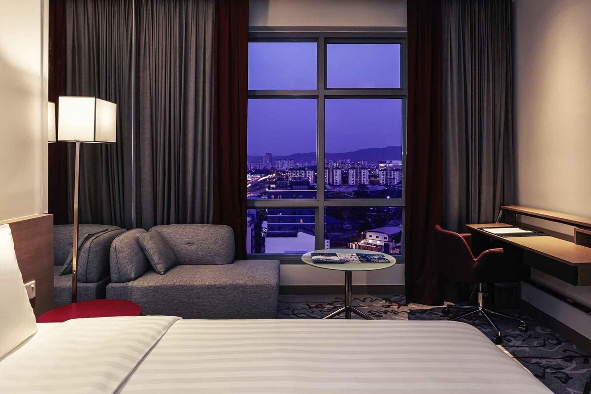 A hotel room with a bed, a sofa, a desk, and a view of a cityscape through a large window.