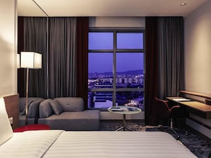 A hotel room with a bed, a sofa, a desk, and a view of a cityscape through a large window. - Mercure Kuala Lumpur Shaw Parade (Kuala Lumpur)