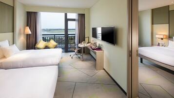 Quarto de hotel com duas camas, uma secretária com cadeira, televisão de tela plana e varanda com vista para a água.