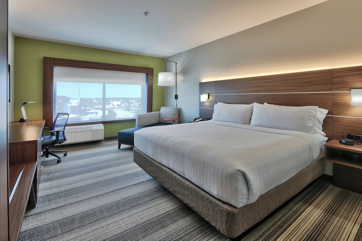 A modern hotel room with a large bed, a desk with a chair, a standing lamp, and a view of the city through the window.