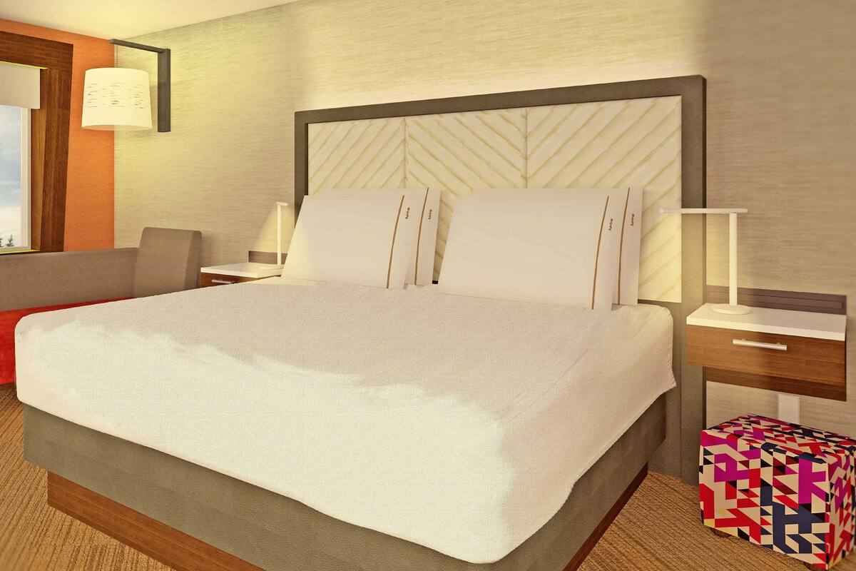 A modern hotel room with a large bed, a red bench, a bedside table with a lamp, and a colorful cube-shaped object on the floor.