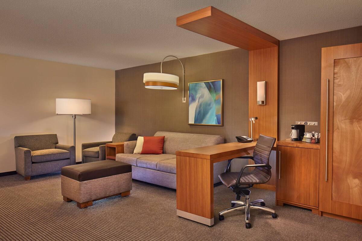 A hotel room with a sofa, armchair, ottoman, desk, and a coffee machine.