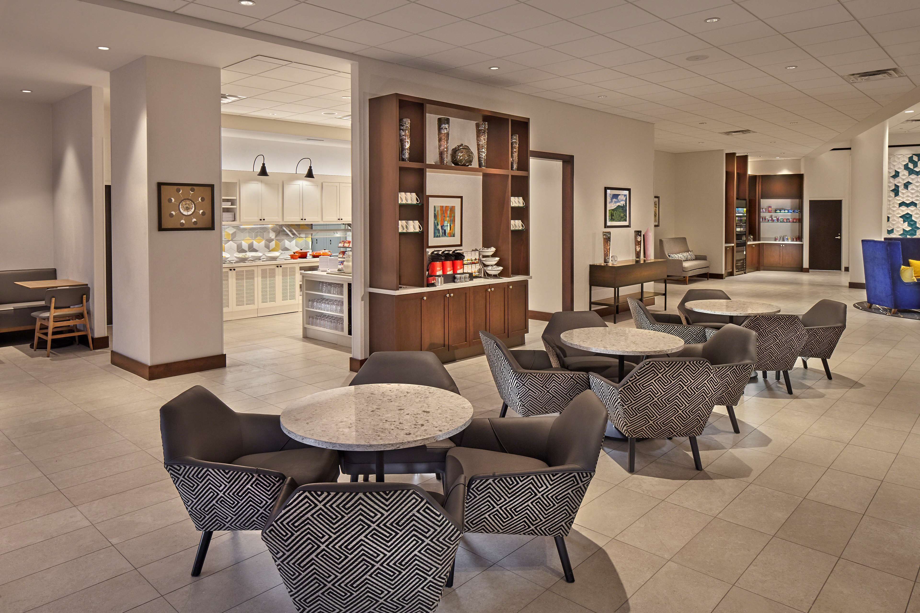 Eine moderne Hotelhalle mit runden Tischen und gemusterten Stühlen, eine Bar mit Regalen voller Flaschen und Blick in die Küche.
