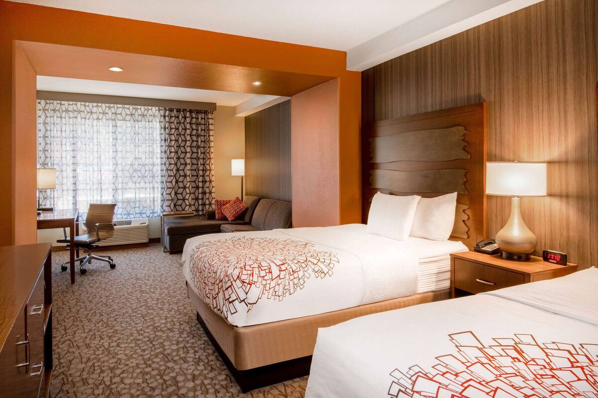 A hotel room with two beds, a desk, a chair, and a small table with a lamp.