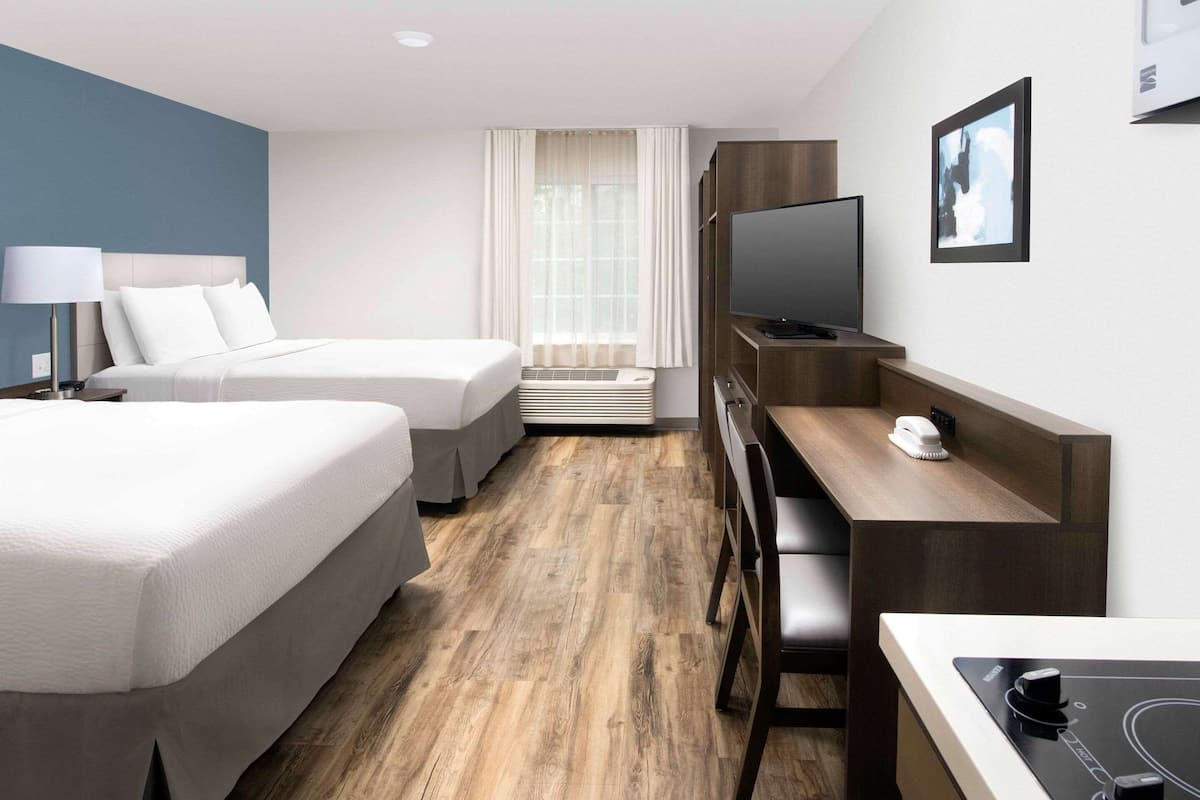 A hotel room with two beds, a desk with a chair, a television, and an air conditioning unit.