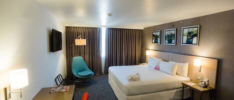 A hotel room with a bed, a desk, a chair, a lamp, a nightstand, and a wall-mounted TV.