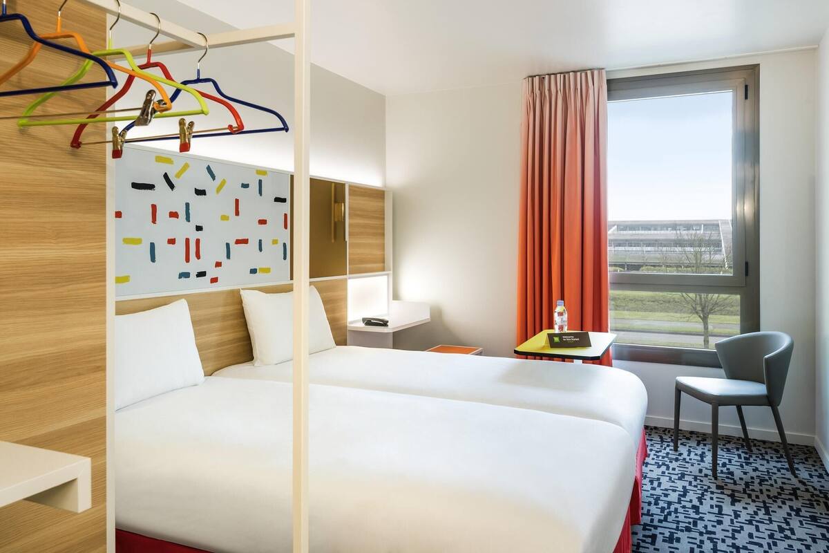 Hotelkamer met een groot bed, een bureau, een stoel, een raam met uitzicht, en een kleurrijk abstract kunstwerk aan de muur.