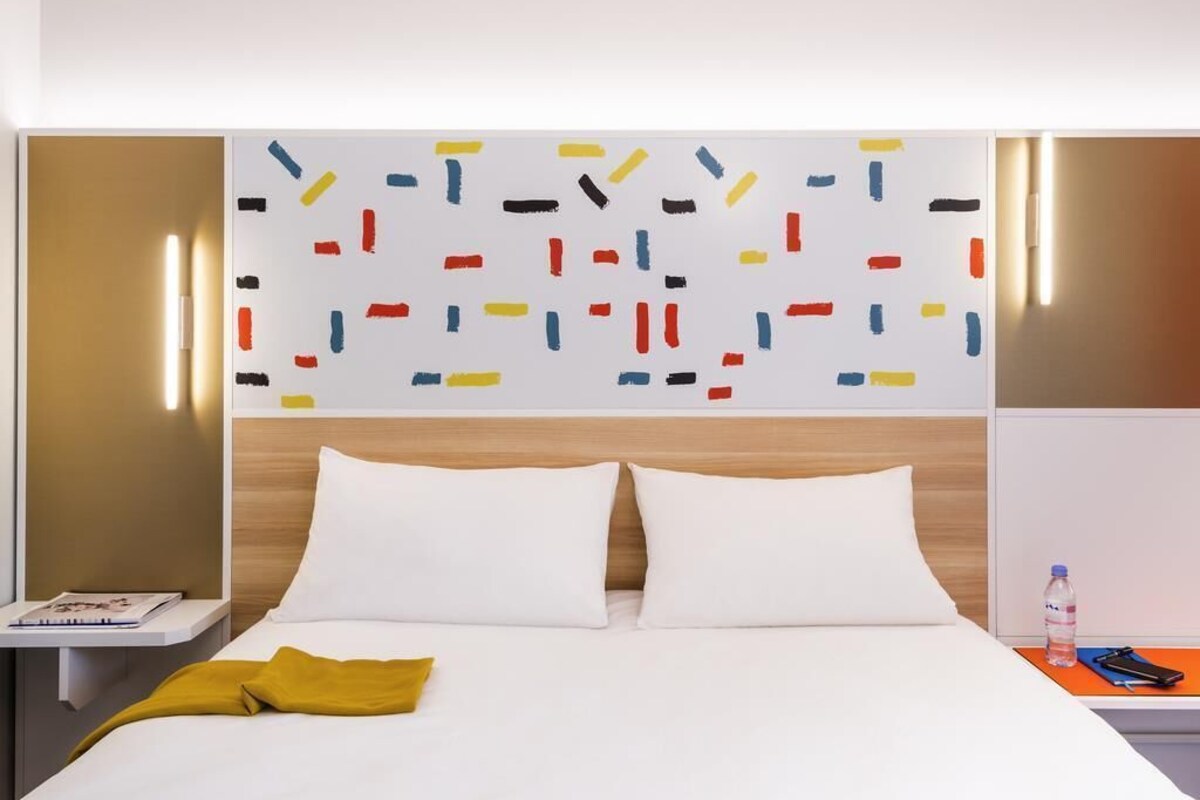 Ein Hotelzimmer mit Bett, weißen Kissen, einer senffarbenen Tagesdecke, einem Nachttisch mit Buch und Flasche sowie einer Wand mit farbenfroher abstrakter Kunst.