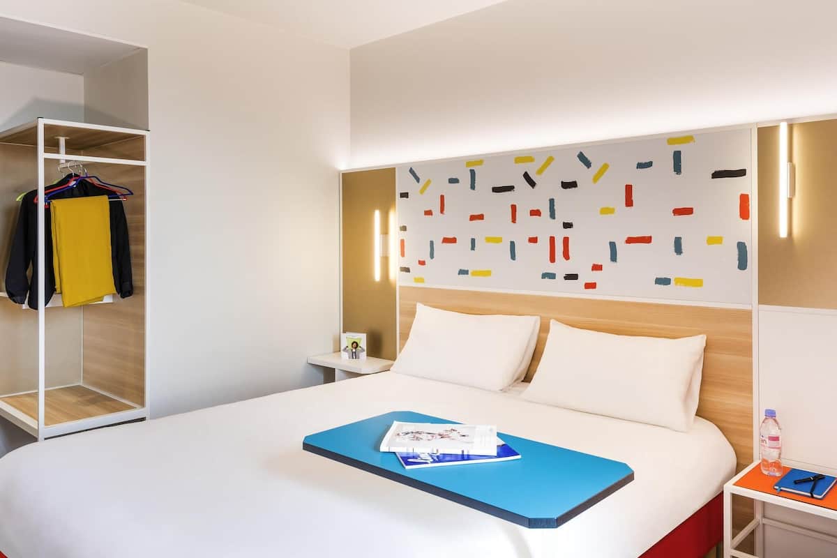Moderni hotellihuone, jossa on siististi pedattu sänky, yöpöytä, jossa on vesipullo, ja värikäs, abstrakti taideteos sängyn yläpuolella.
