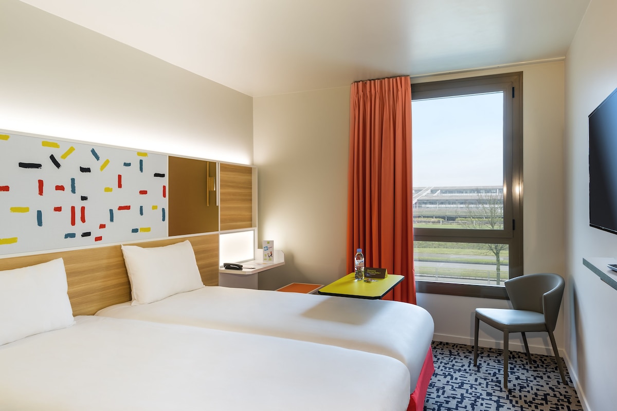 Ein Hotelzimmer mit einem großen Bett, einem kleinen Tisch mit Flasche und Glas, einem Sessel und einem Fenster mit Vorhängen.