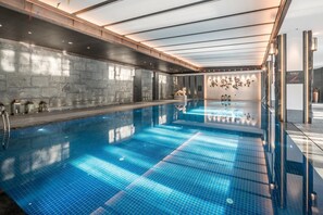 Una amplia piscina interior con suelo de baldosas azules brillantes, pared de piedra en un lateral y techo de madera con tragaluces.