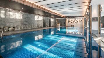 A large indoor swimming pool with a clear blue tiled floor, a stone wall on one side, and a wooden ceiling with skylights.