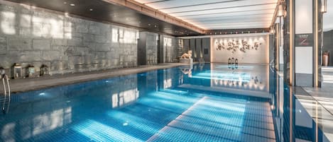 A large indoor swimming pool with a clear blue tiled floor, a stone wall on one side, and a wooden ceiling with skylights.