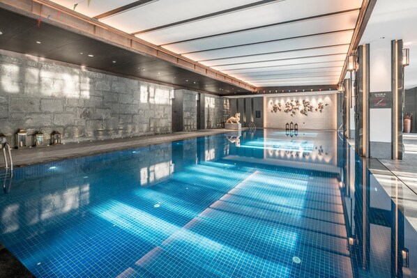 A large indoor swimming pool with a clear blue tiled floor, a stone wall on one side, and a wooden ceiling with skylights.