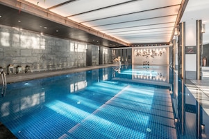 A large indoor swimming pool with a clear blue tiled floor, a stone wall on one side, and a wooden ceiling with skylights.