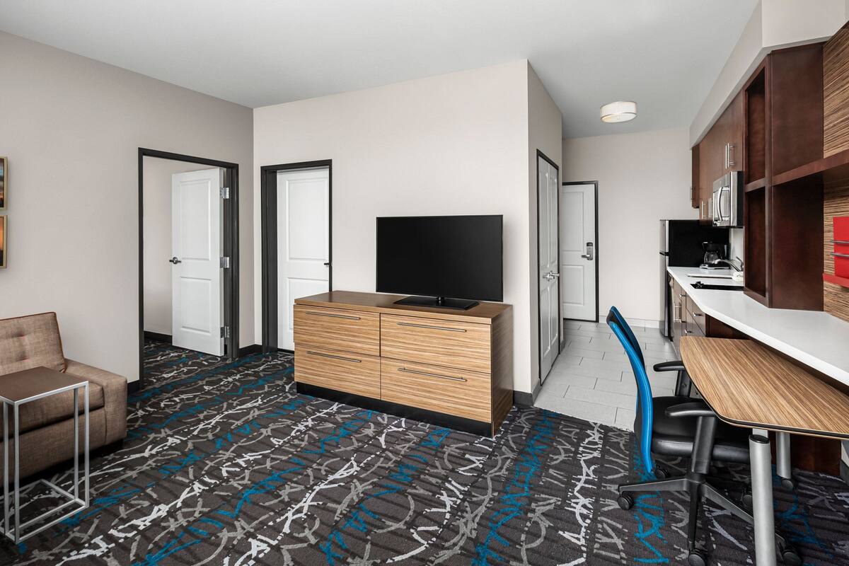 Habitación de hotel con cómoda de madera, televisión de pantalla plana, escritorio con silla, kitchenette y alfombra estampada.