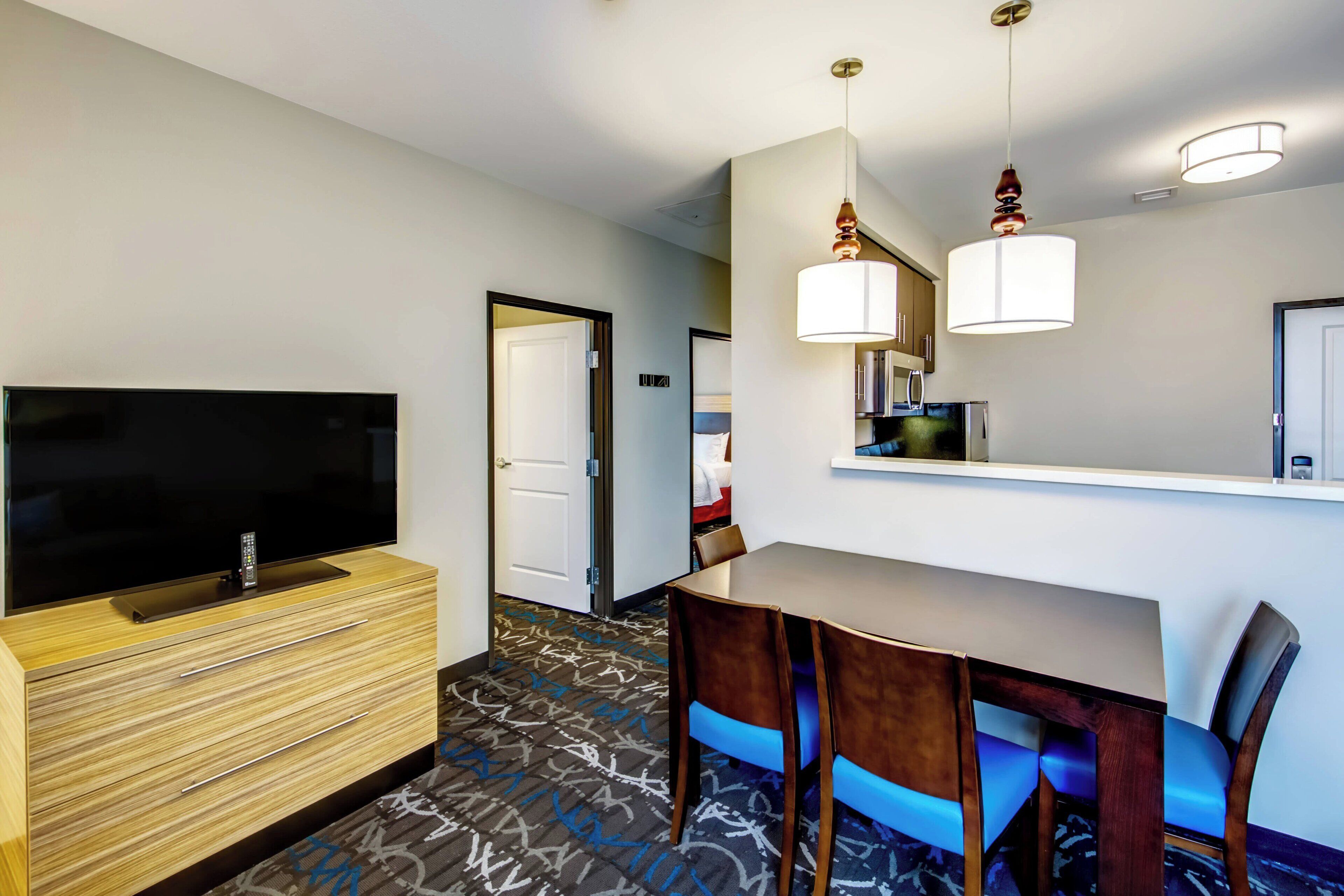 A hotel room with a flat-screen TV, wooden dresser, dining table with chairs, and a patterned carpet.
