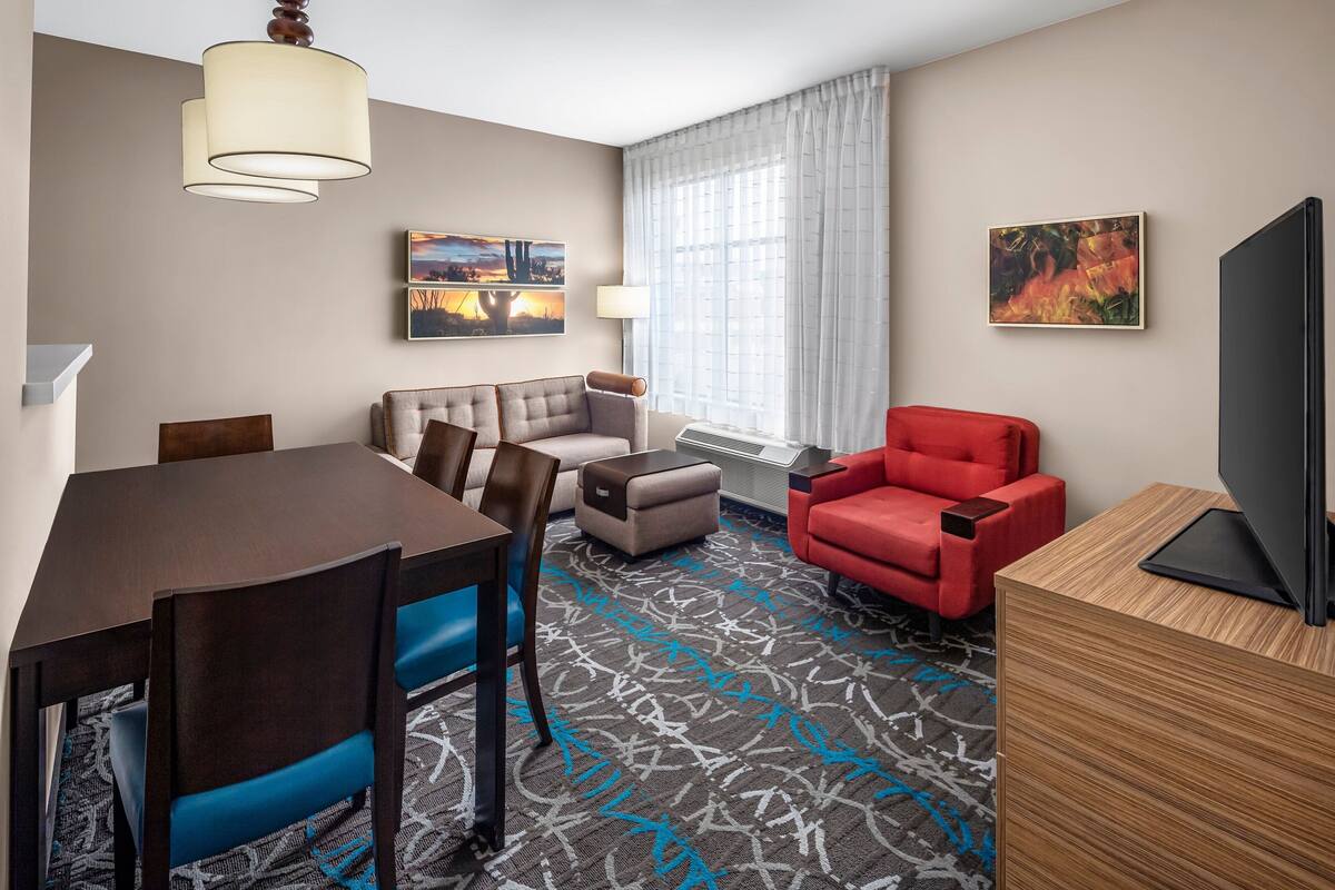 Habitación de hotel con una mesa de comedor, sillas, un sofá, un sillón rojo, un televisor y obras de arte en las paredes.