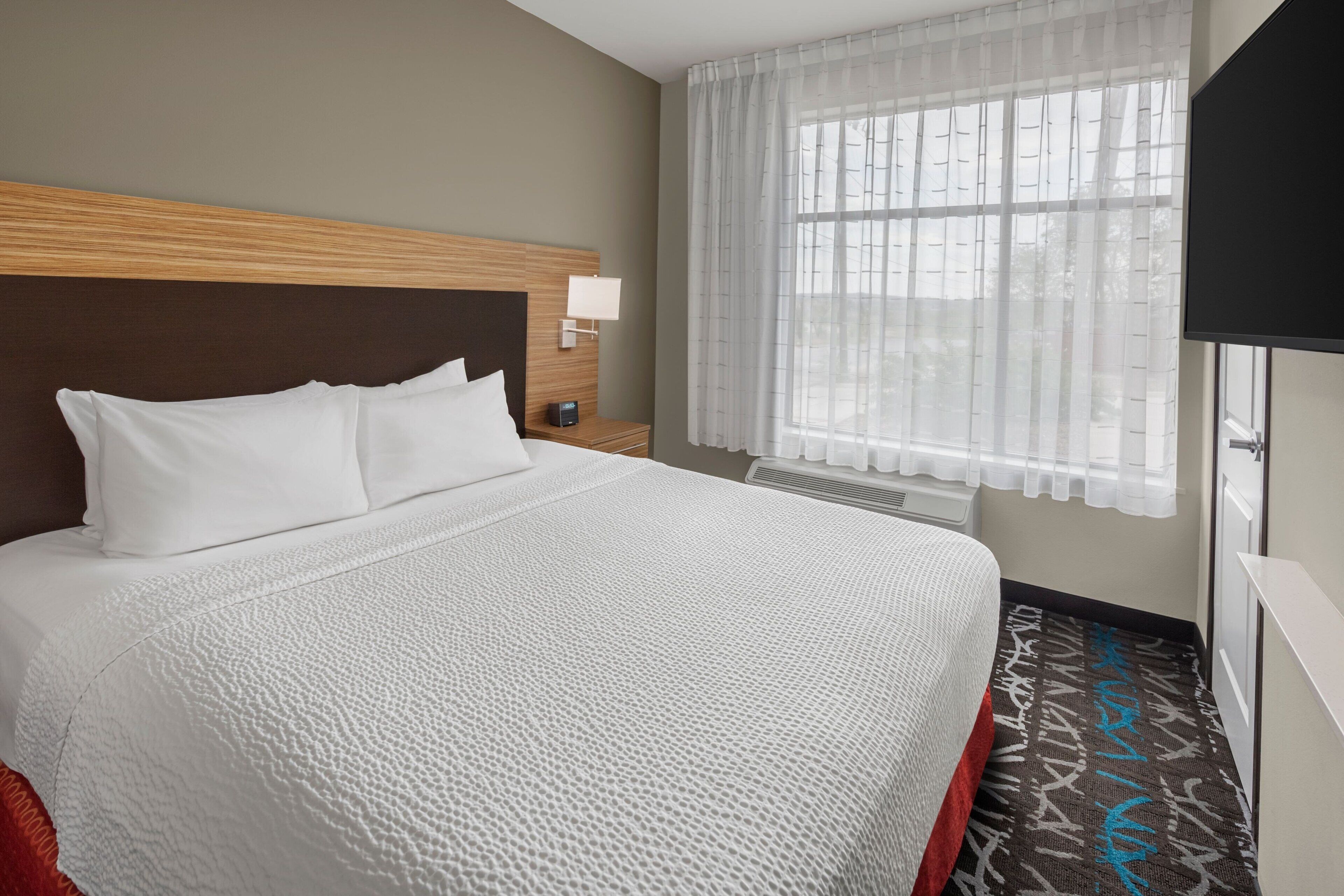 A hotel room with a large bed, a bedside table with a lamp, a window with sheer curtains, and a flat-screen TV mounted on the wall.