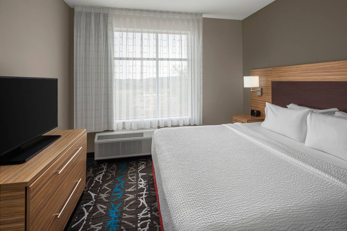Habitación de hotel con cama, televisor sobre una cómoda de madera, ventana con cortinas translúcidas y mesita de noche con lámpara.