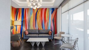 Une chambre d’hôtel moderne avec un mur décoré d’une œuvre abstraite aux couleurs vives, un canapé gris, une table basse blanche et une cloison en verre offrant une vue dégagée.
