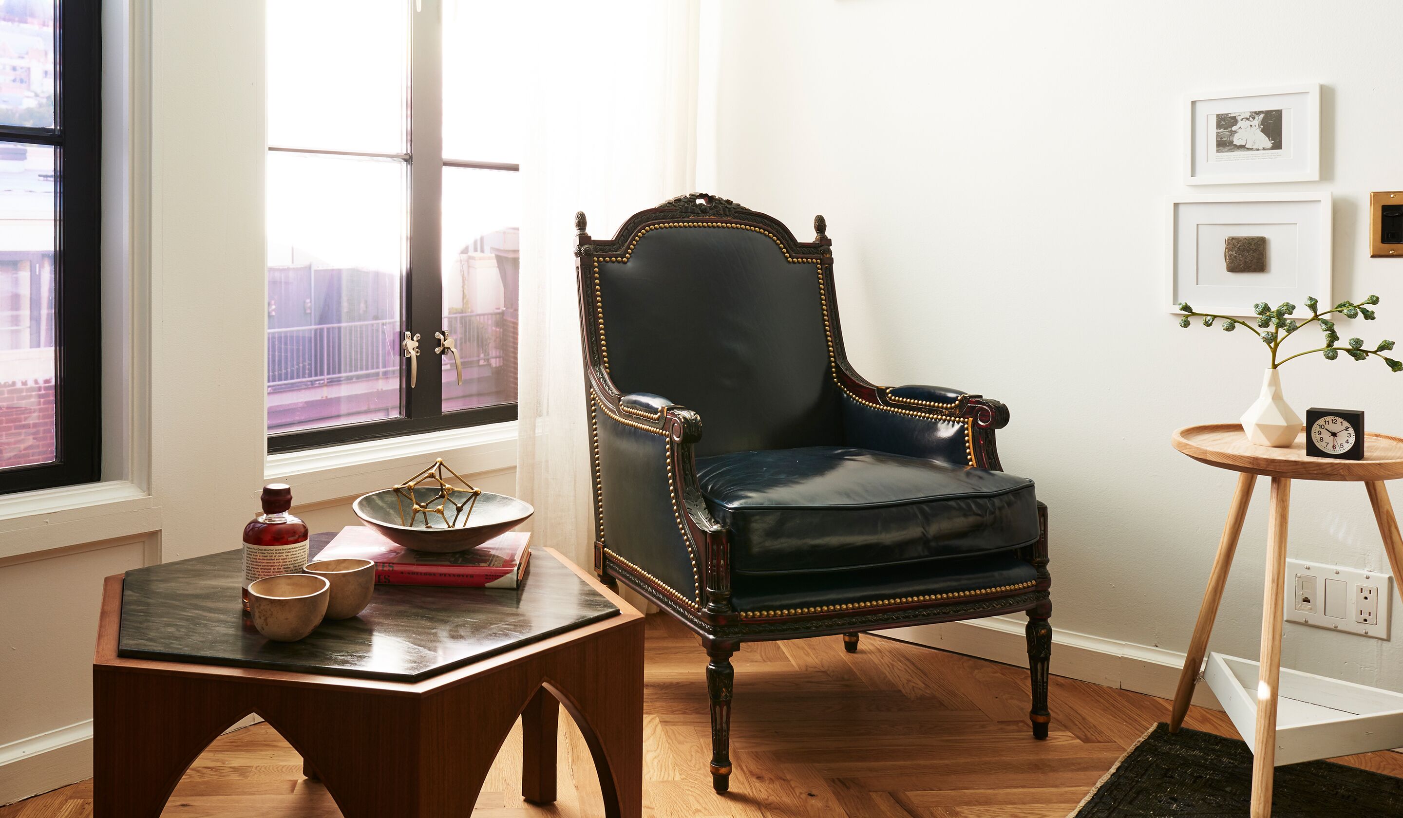 A room with a dark blue upholstered chair, a small wooden table with a bowl and books, a vase with a plant, and a framed picture on the wall.