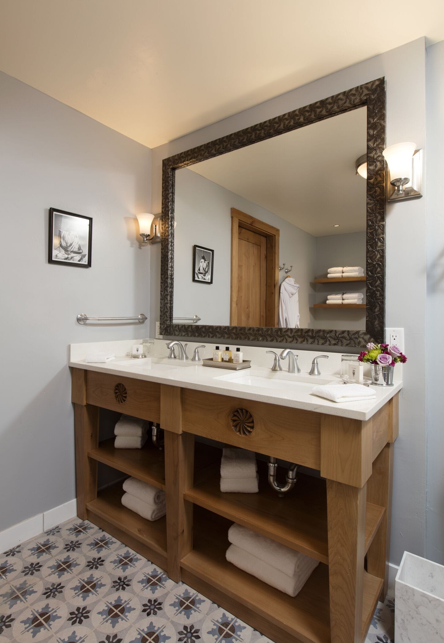 A bathroom with a large mirror, two sinks, wooden vanity, towel racks, and a patterned floor.