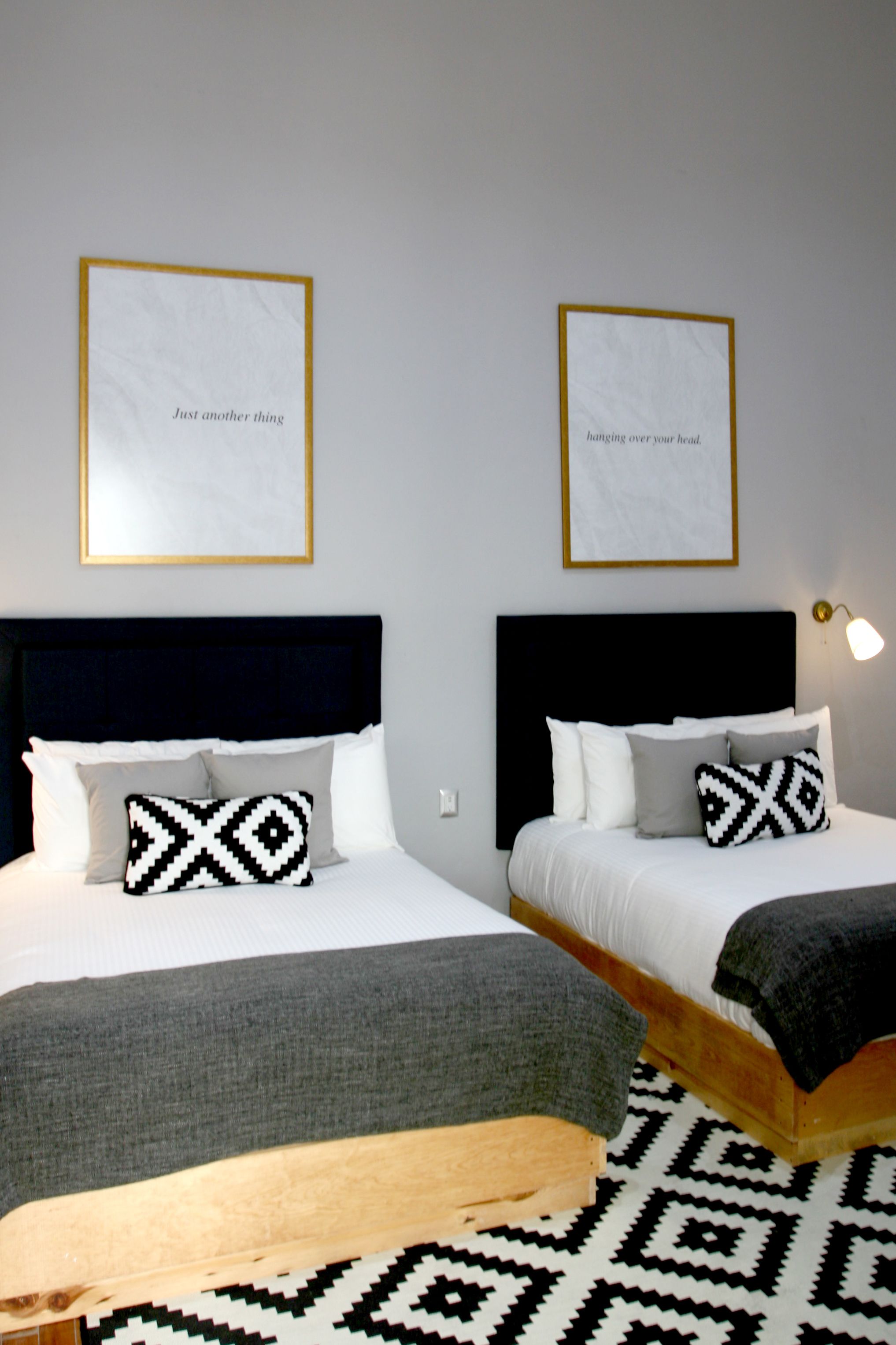 Habitación de hotel con dos camas, cada una con cabecero negro, ropa de cama blanca y almohadas estampadas. Hay cuadros enmarcados en la pared y una lámpara de noche.