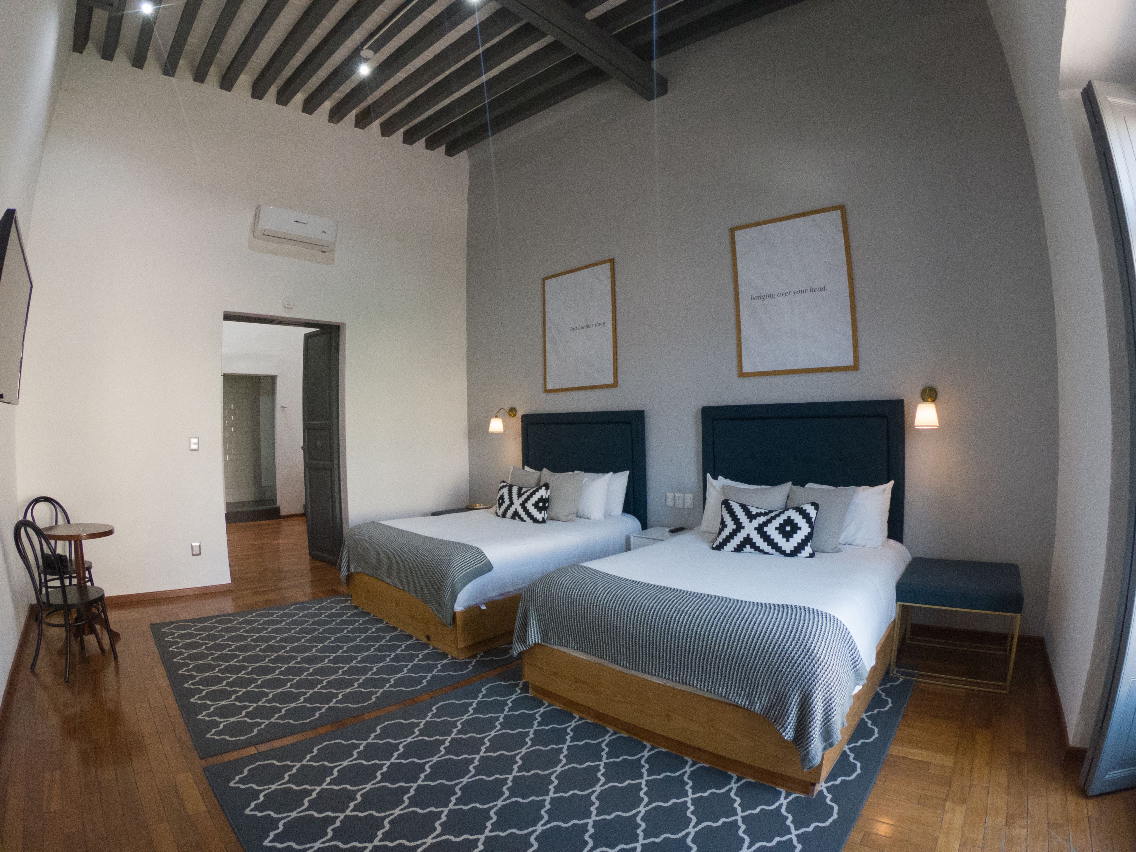 Habitación de hotel con dos camas, una mesita y una silla. Se observan cuadros en la pared y una lámpara en el techo.