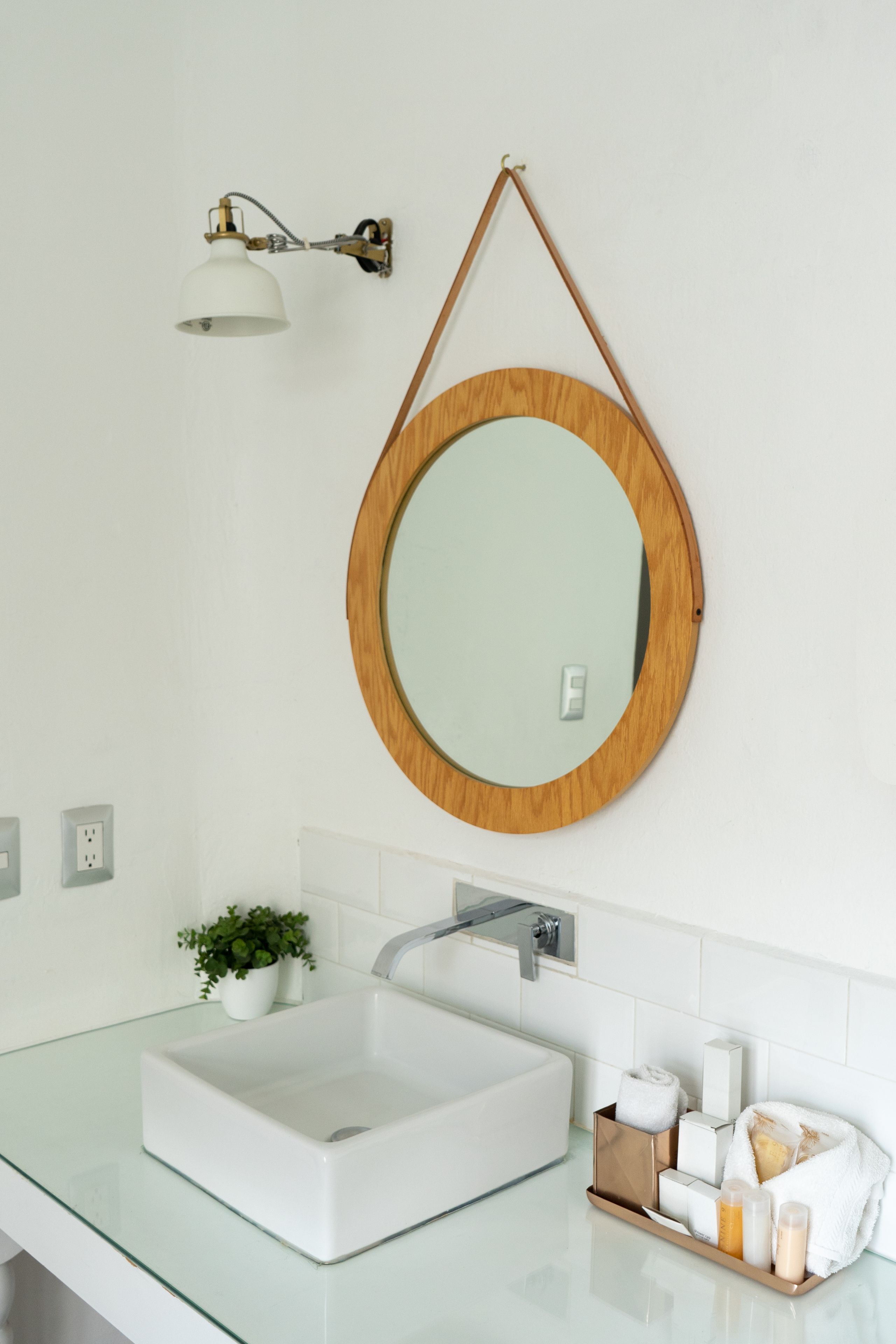 A bathroom with a white sink, a round mirror with a wooden frame, a wall-mounted light fixture, and a small potted plant.