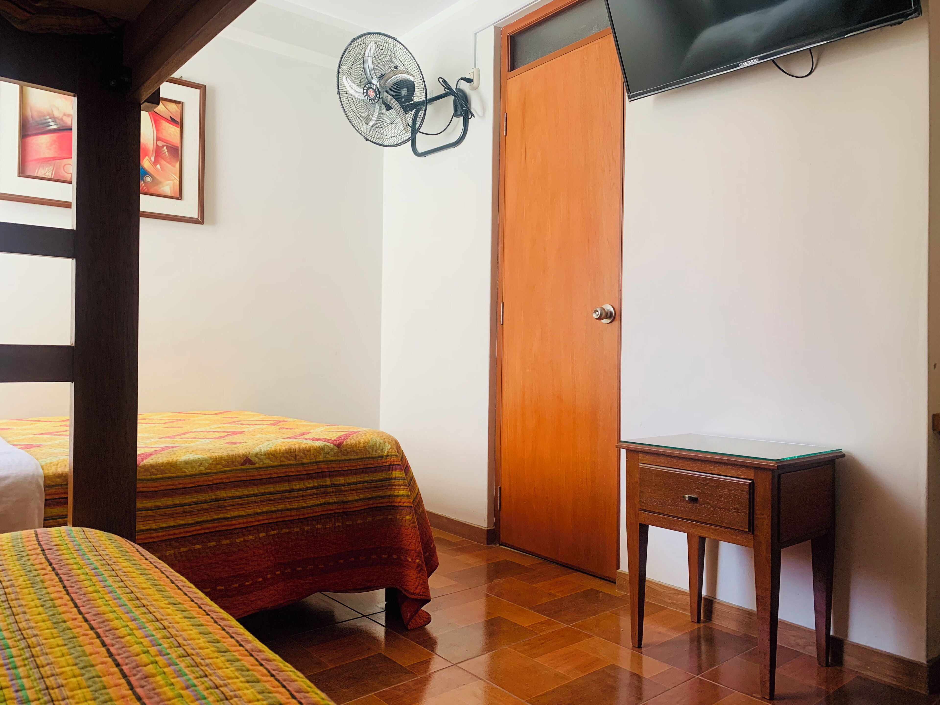 A bedroom with a bed, a wooden door, a small table, a fan, and a TV mounted on the wall.