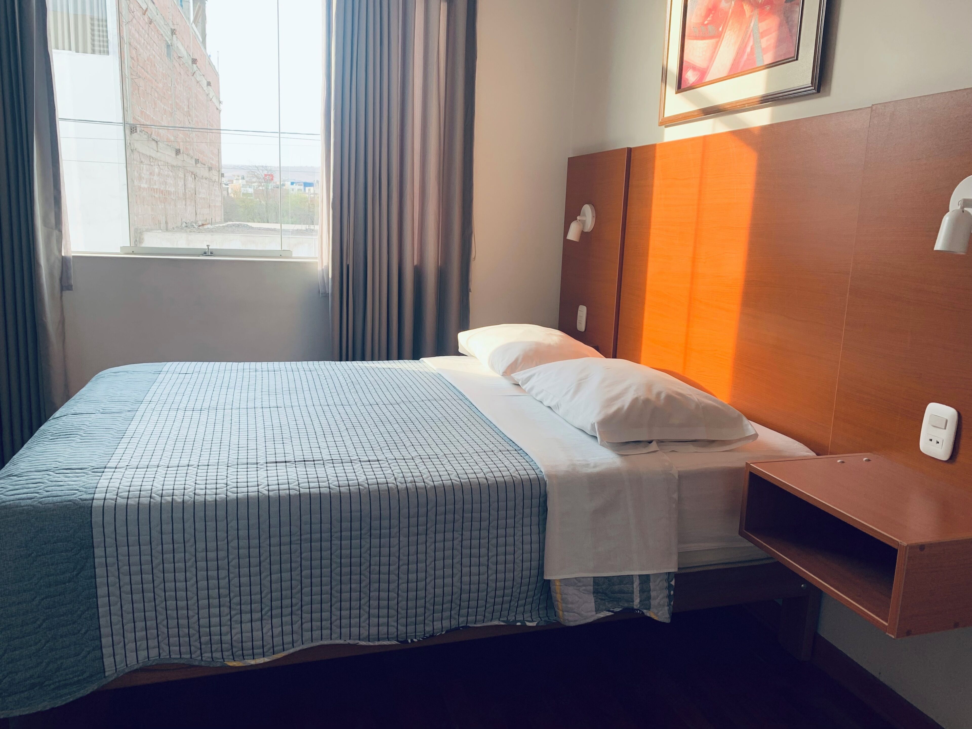 Habitación de hotel con cama, mesita de noche, ventana con cortinas, lámpara de pared y un cuadro enmarcado.