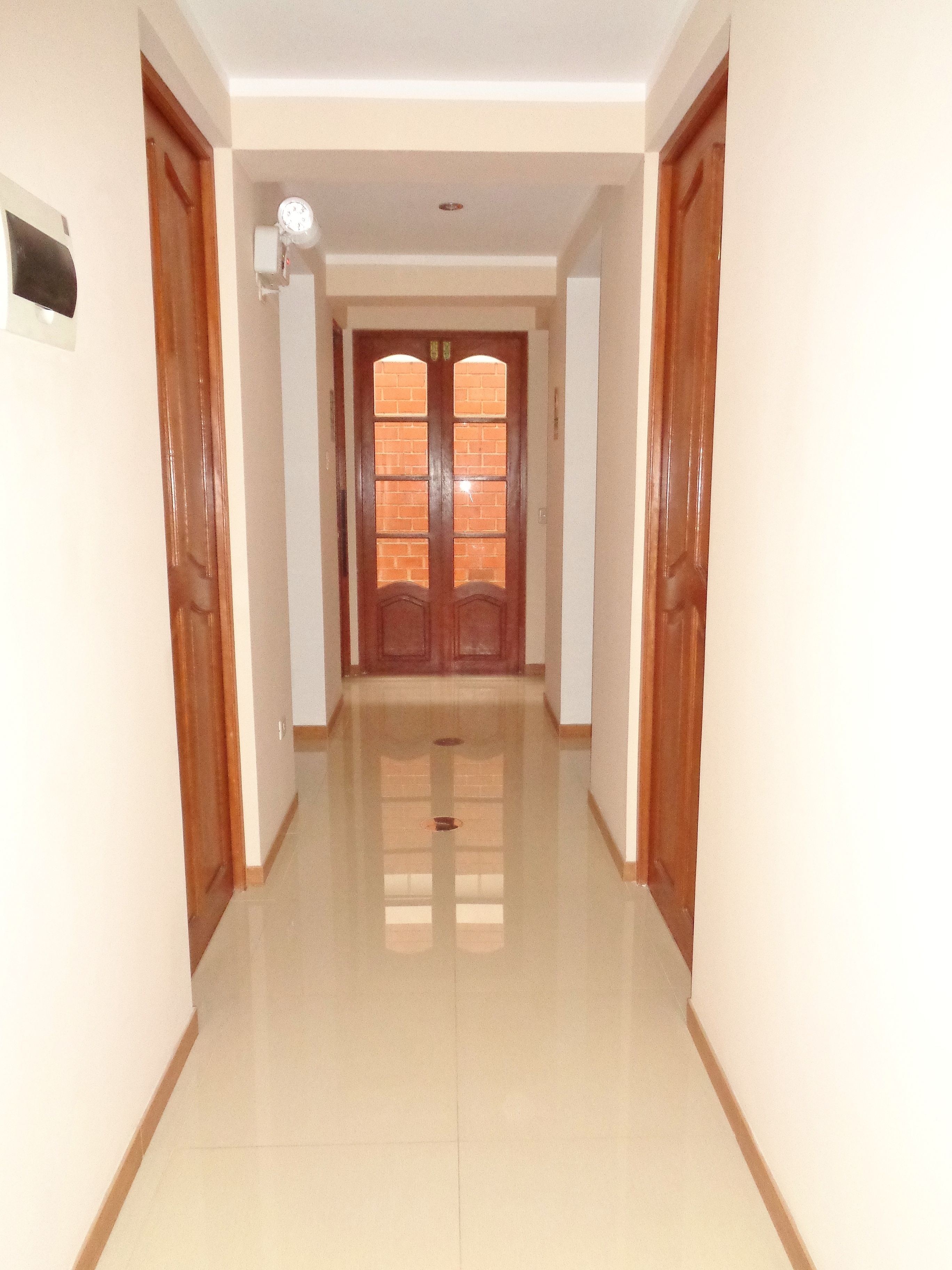 Un pasillo con una puerta de madera al final, paredes blancas y piso de baldosas.