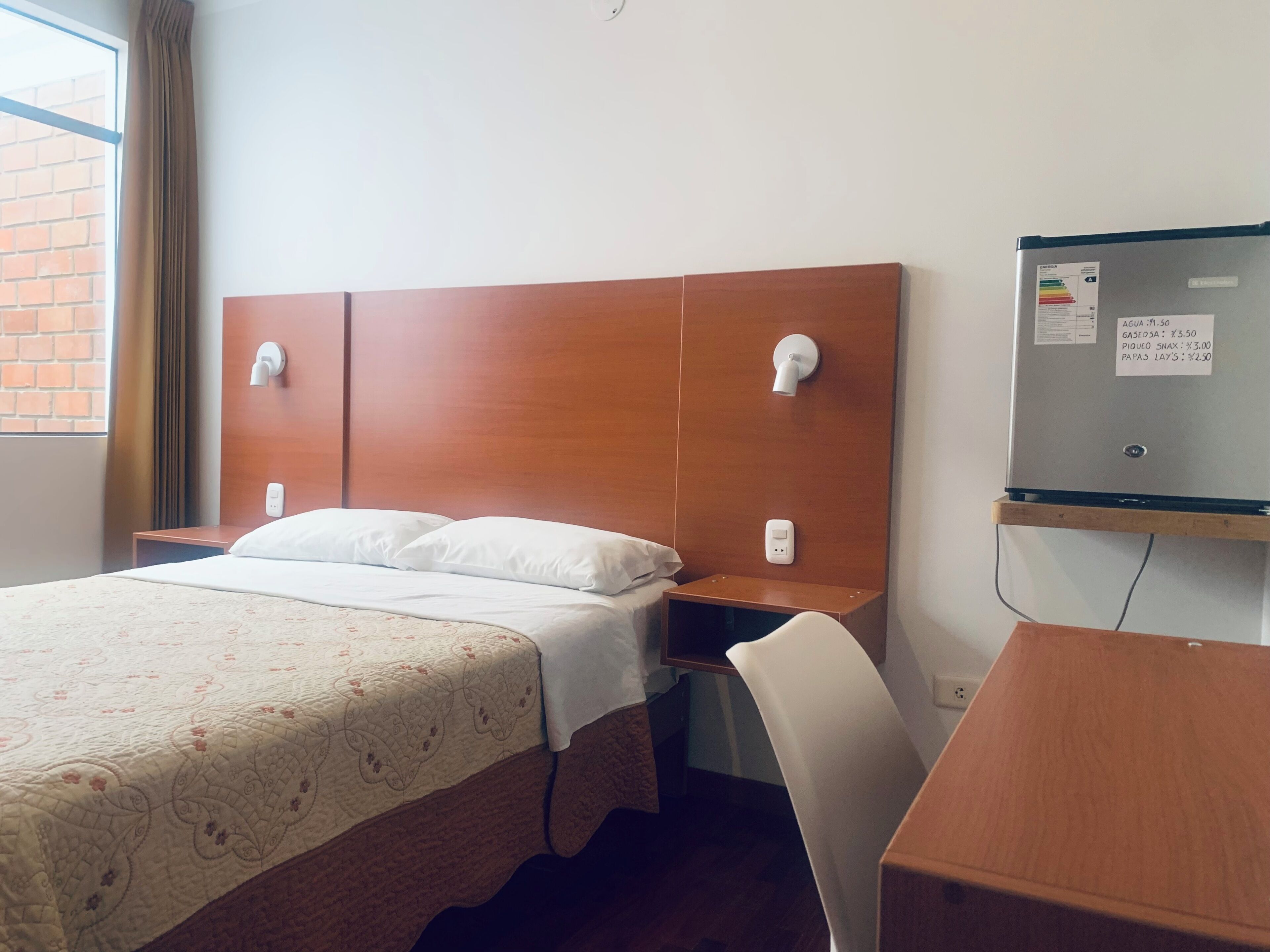Quarto de hotel com cama, mesa de cabeceira, cadeira e frigorífico.