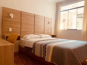 A hotel room with a wooden headboard, a bed with a patterned blanket, a desk with a chair, and a window with curtains.